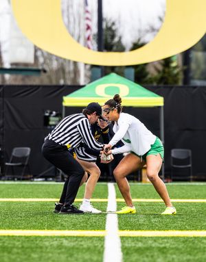 A women's lacrosse player faces off against an opponent with a referee on the University of Oregon turf field.