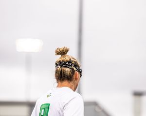 Female soccer player with hair bun and black headband wearing green and white jersey viewed from behind.