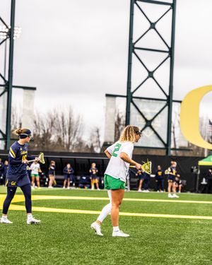 Two women's lacrosse players compete on a turf field with the Oregon Ducks logo visible in the background.
