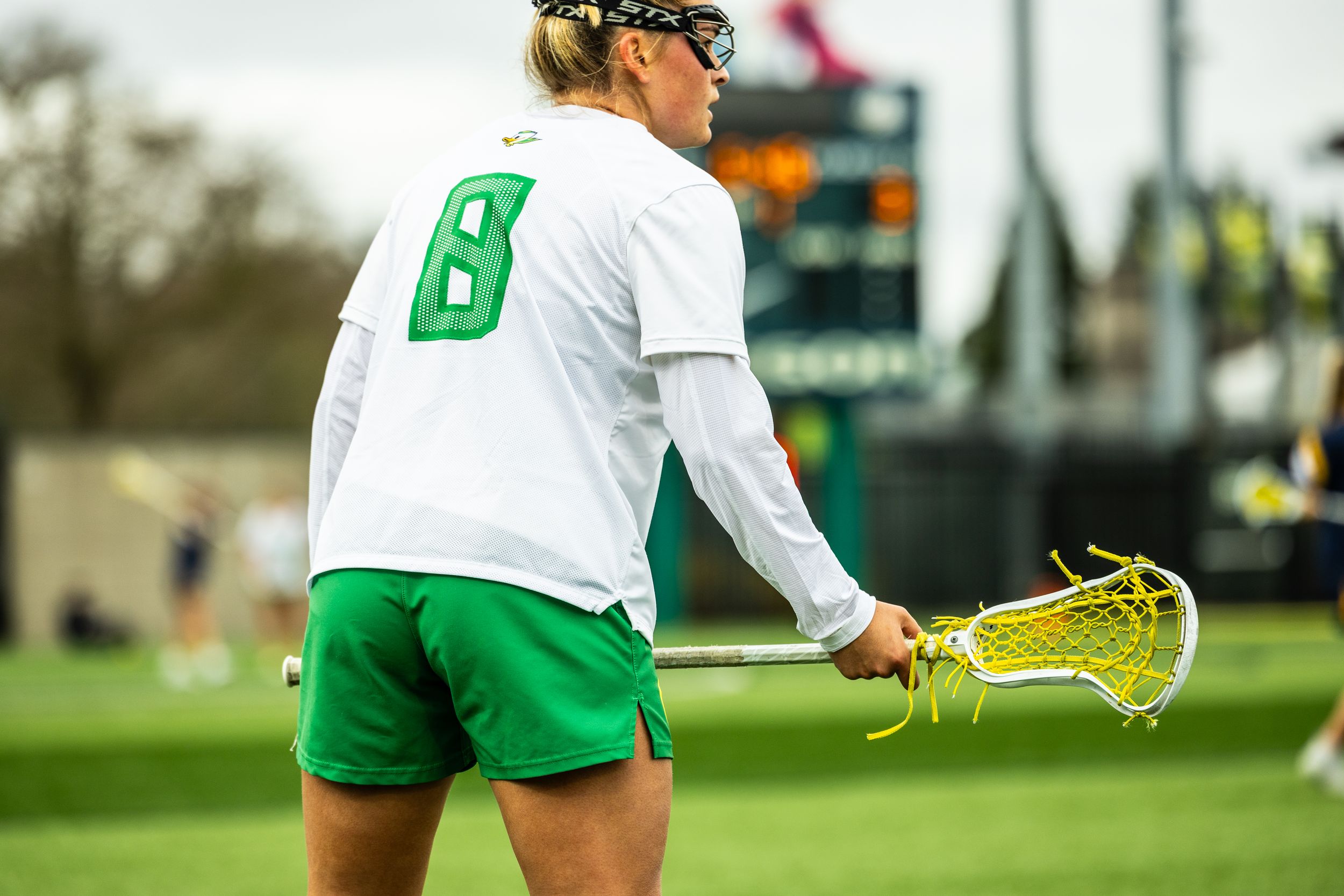 Oregon Ducks female lacrosse player wearing green shorts and white jersey number 8, holding a lacrosse stick on the field.