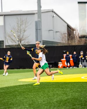 Two female lacrosse players compete on a turf field, one in green and one in navy, during an outdoor collegiate game.
