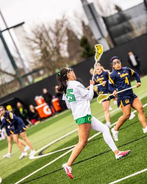 Female lacrosse player in green uniform winds up to shoot during a college women's lacrosse game on turf field.
