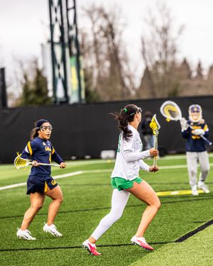 Women's lacrosse players compete on a turf field, with Cal Bears players defending against an Oregon player.