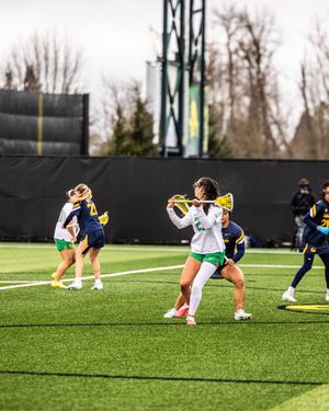 Women's lacrosse player in white Oregon uniform cradles ball while defended by opponents in navy uniforms on turf field.