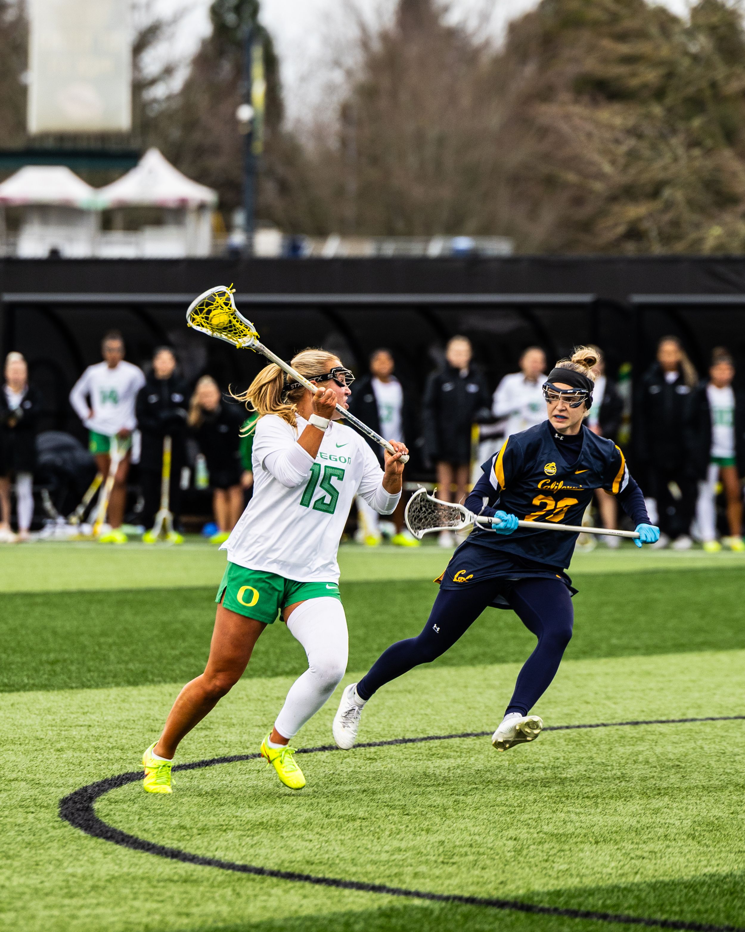 Two women's lacrosse players compete on a turf field, one in green Oregon uniform, the other in Cal Berkeley navy.