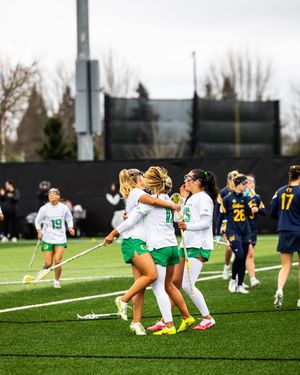 Women's lacrosse players in green uniforms celebrate on turf field during competitive college game.