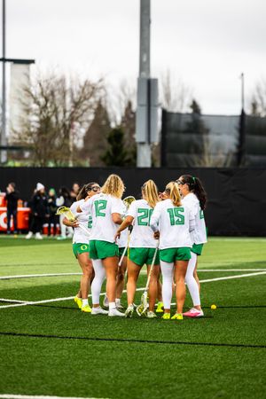 Oregon Ducks women's lacrosse team huddles on turf field in white and green uniforms during a match.