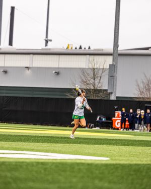 Oregon Ducks female lacrosse player running on turf field, holding stick overhead during a college game.