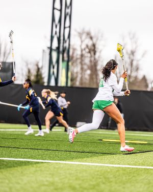 Female lacrosse player in green shorts sprints on turf field during competitive game.