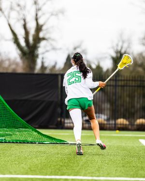 Female lacrosse player #25 in green and white uniform running on turf field holding a lacrosse stick near the goal net.