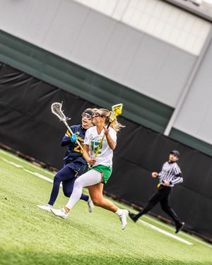 Female lacrosse players in green and navy uniforms battle for possession during an indoor turf game.