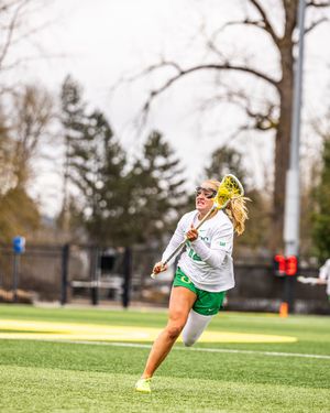 Female lacrosse player in green and white uniform running with stick during outdoor game on turf field.