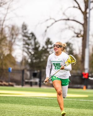 Oregon Ducks female lacrosse player #15 running with stick on turf field in white and green uniform.