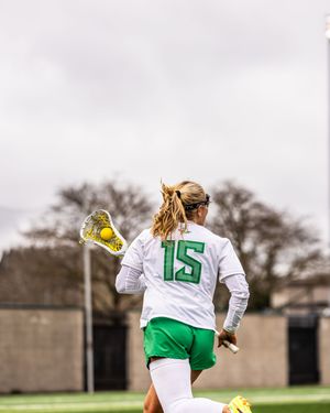Female lacrosse player wearing number 15 white and green uniform running with stick and ball on outdoor field.