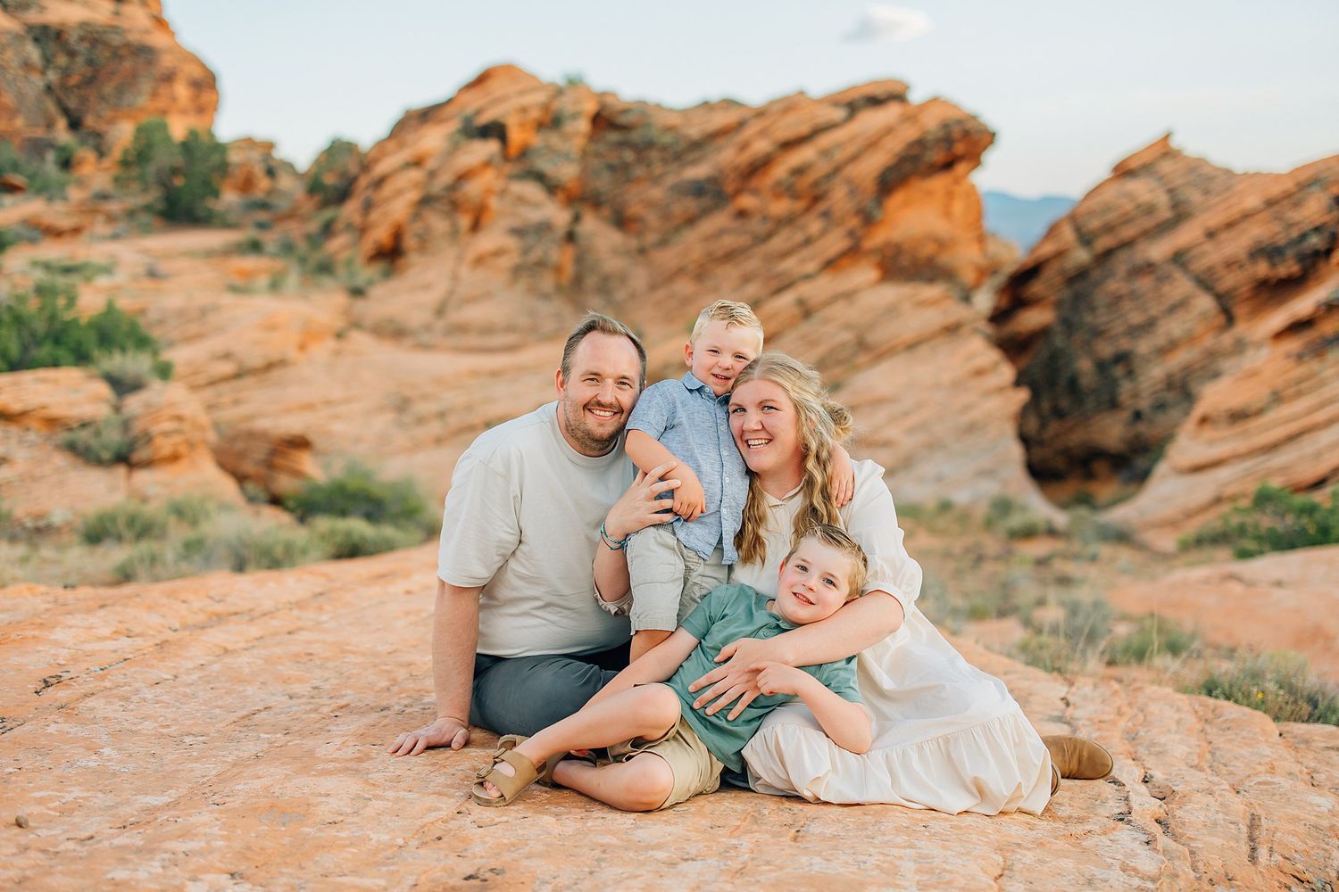 Family of four smiling together on red rock formations in a desert landscape during golden hour.