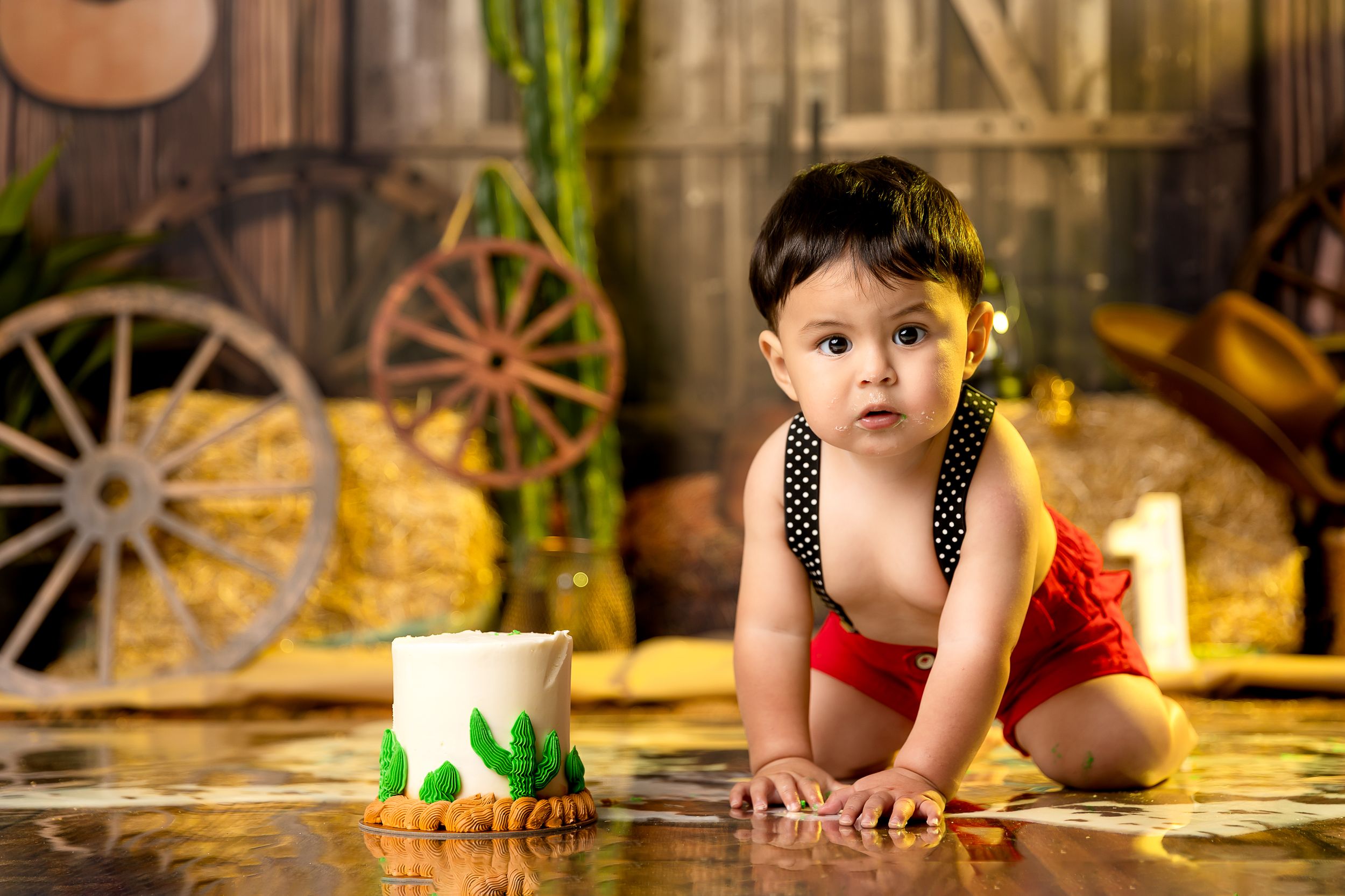 A baby in red bloomers and polka dot suspenders crawls near a small cake in a rustic barn-themed photo setting.