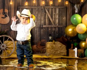 Toddler in cowboy hat and jeans poses in rustic barn setting with balloons and hay bales for first birthday.