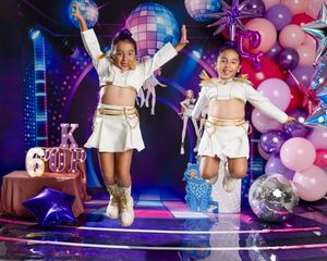 Two girls in matching white and gold outfits jump joyfully at a disco-themed birthday party with colorful balloons and a mirror ball.