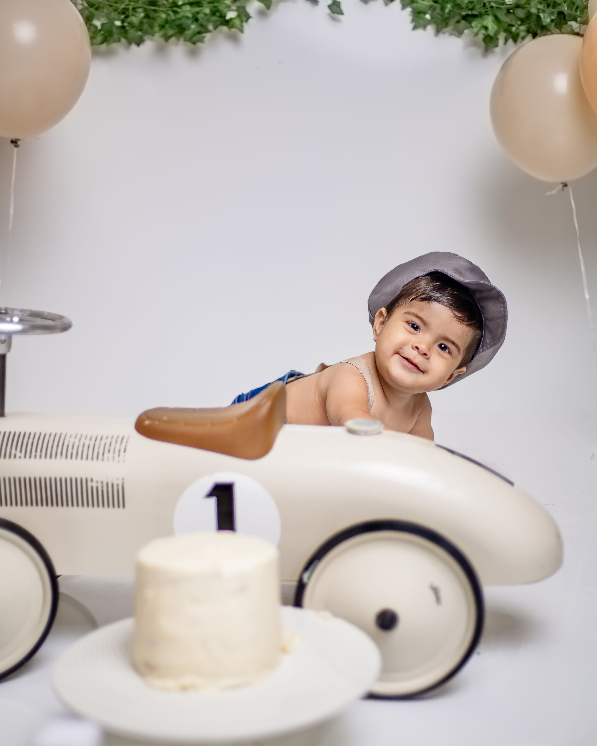 Baby boy in a hat sits in a vintage white race car toy for his first birthday cake smash photo session.