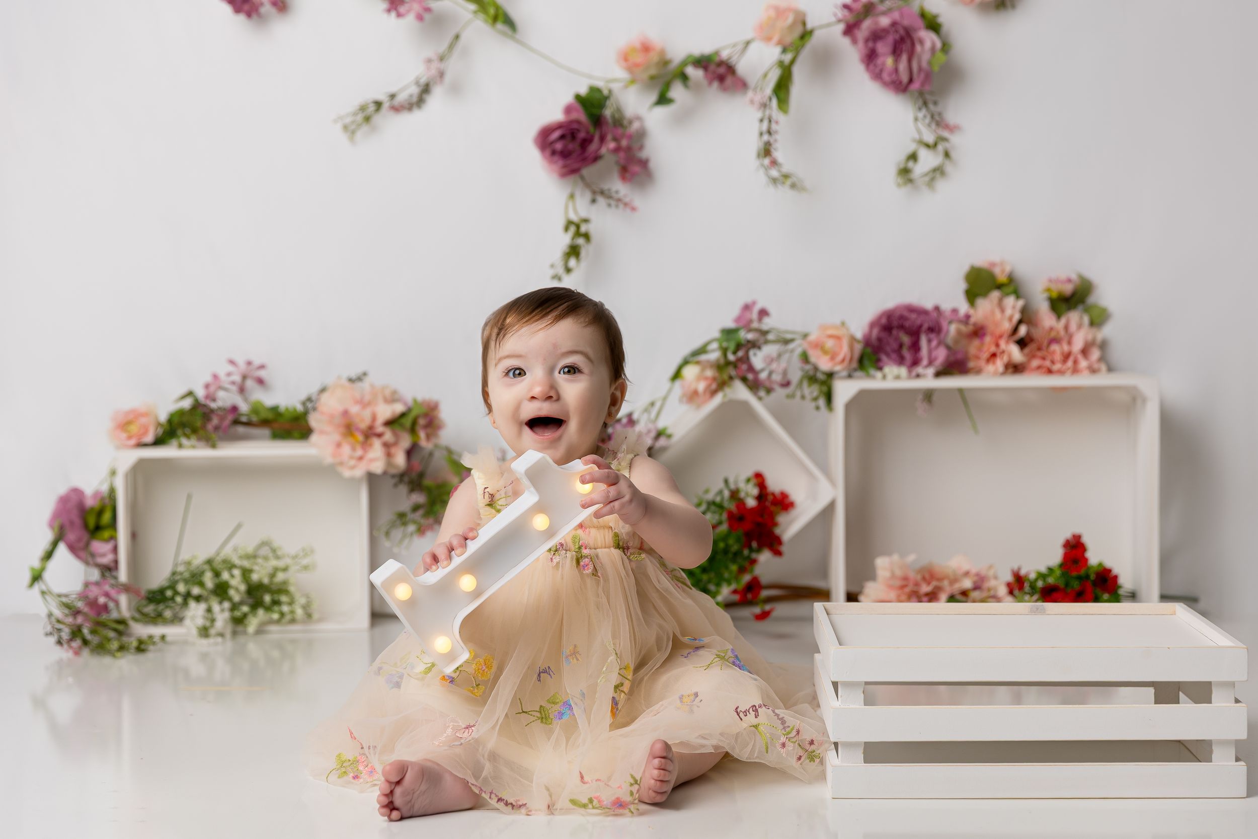 Baby girl in yellow floral dress laughing during first birthday cake smash photo session with flower wall backdrop.