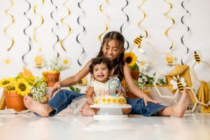Older sibling hugging baby at bee-themed first birthday cake smash with sunflowers and yellow decorations.