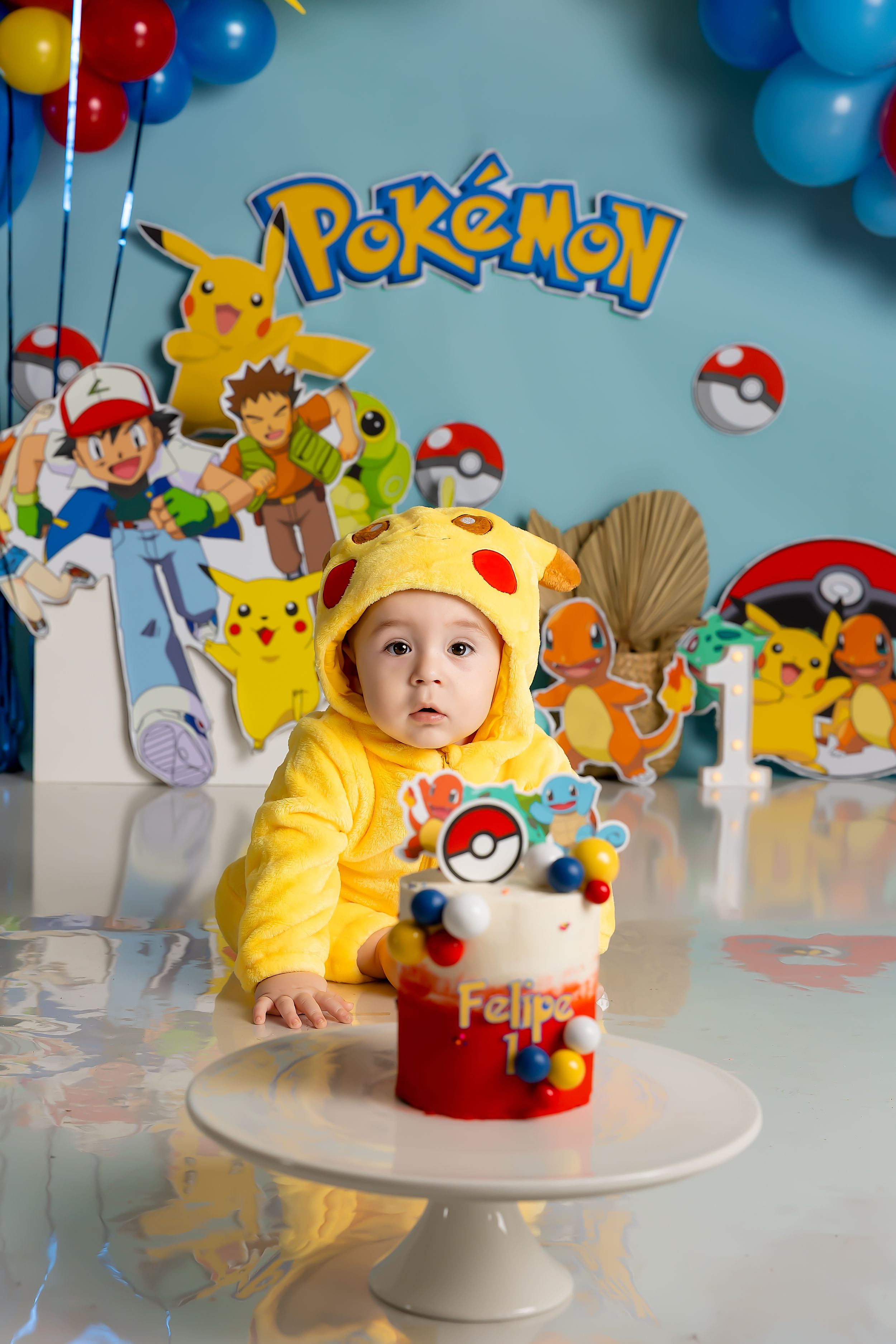 Baby in a yellow Pikachu onesie sits beside a small Pokémon smash cake at a first birthday photoshoot with a Pokémon backdrop.