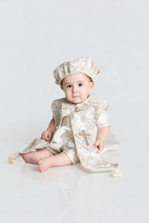 Baby boy sitting in an ivory and gold brocade christening outfit with matching cap and a cross embroidered on the vest.