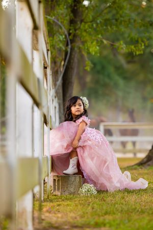 Young girl in pink ball gown and white boots sits beside a fence in a lush outdoor setting.