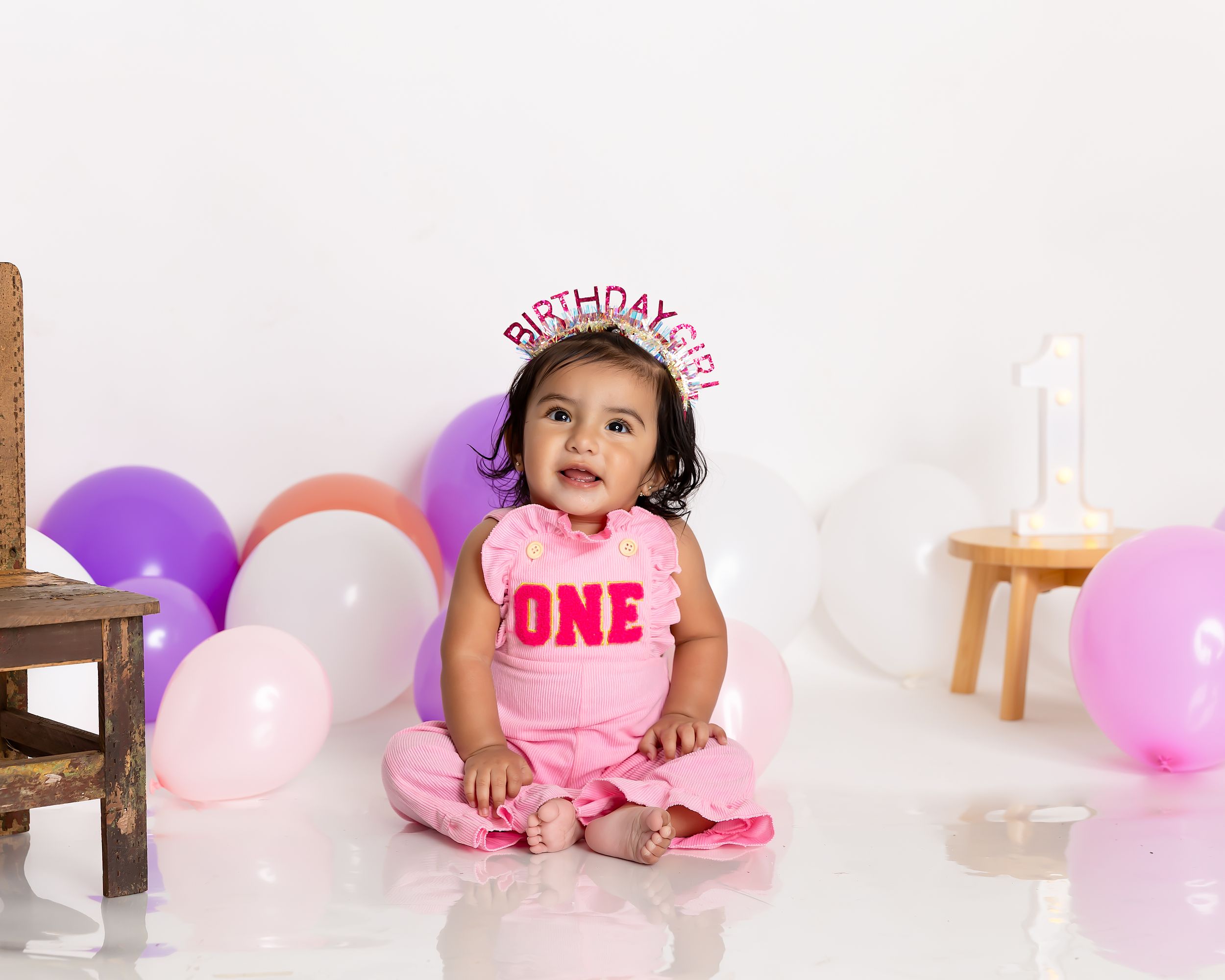Baby girl in a pink "ONE" outfit and birthday girl crown sits among pastel balloons at her first birthday photoshoot.