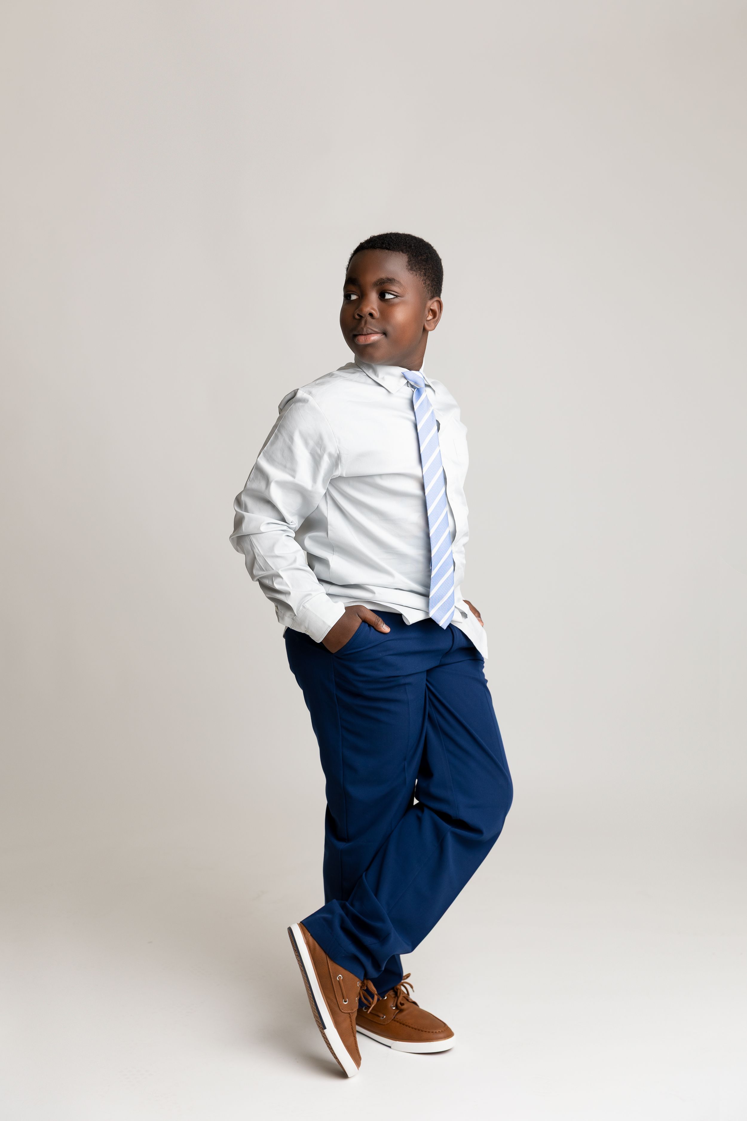 A young Black boy in a white dress shirt, blue tie, navy pants, and brown shoes poses confidently against a gray background.