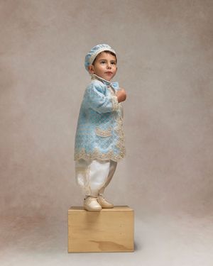 Toddler in blue lace coat and matching bonnet stands on wooden box in a professional portrait photo.