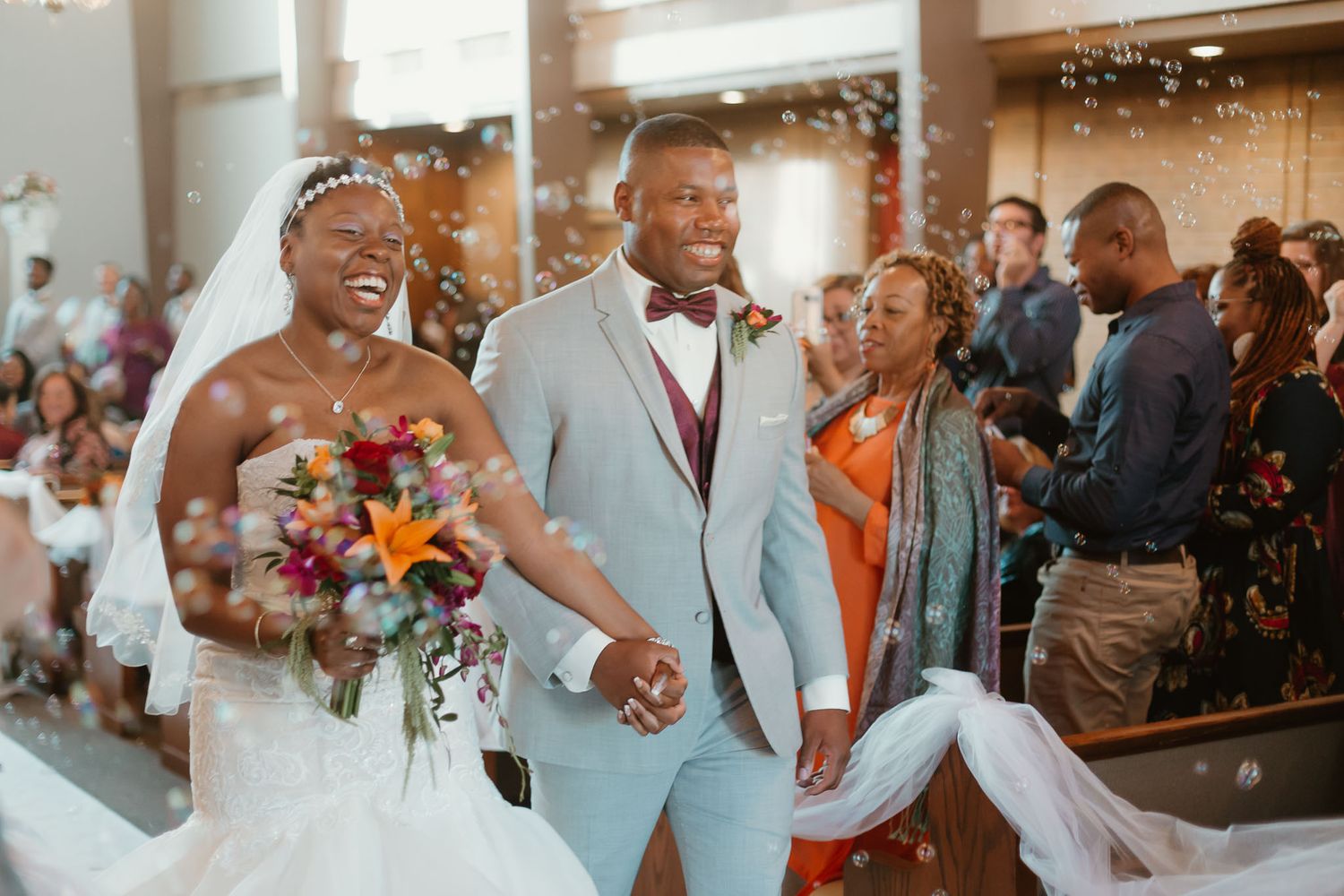 Joyful bride and groom walking down church aisle with colorful bouquet and bubbles.