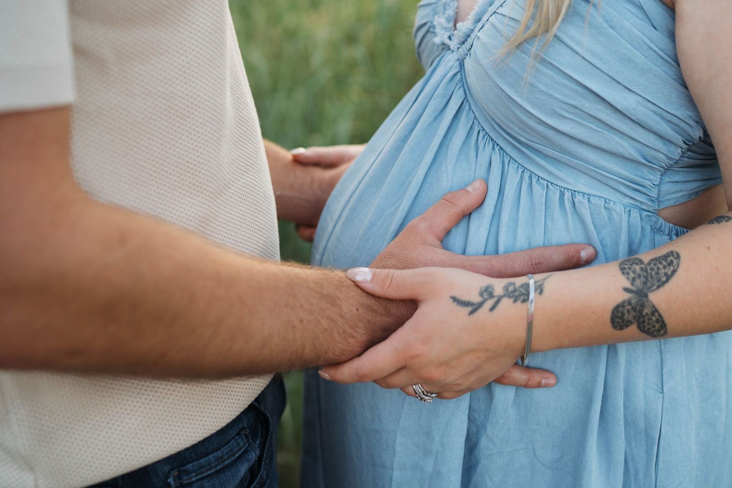 Couple tenderly holding pregnant belly in blue dress outdoors, woman with butterfly tattoo on arm.