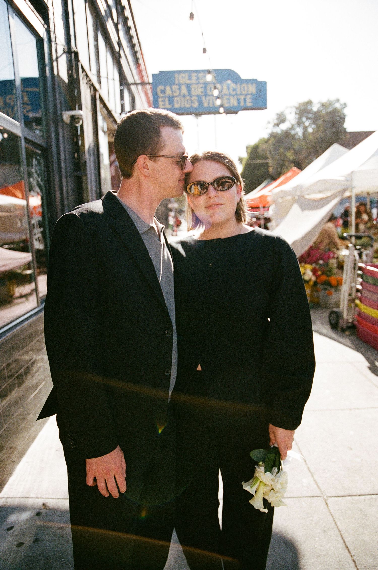 A couple in black outfits shares a tender moment on a sunny street, woman holding white flowers near a colorful market.