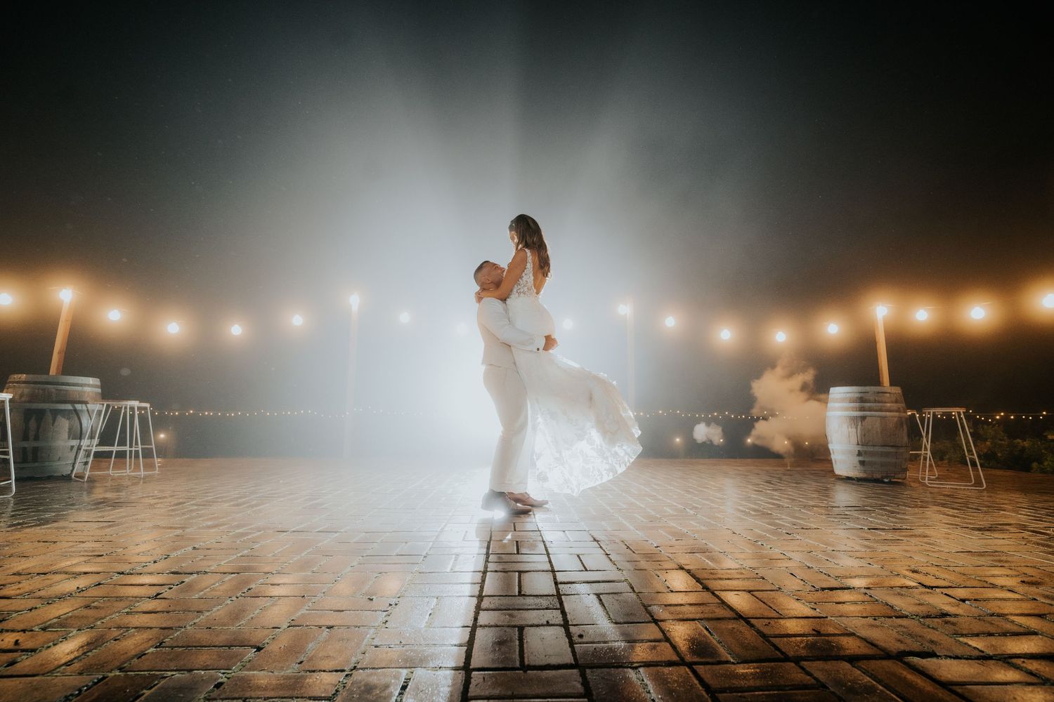 Groom lifting bride on an outdoor dance floor under string lights with misty atmospheric lighting.