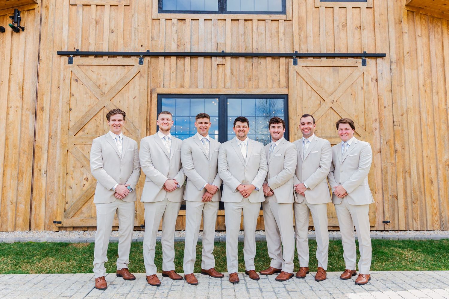 Seven groomsmen in matching tan suits pose together in front of a rustic wooden barn on a wedding day.