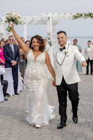 Joyful newlyweds walk down outdoor aisle, bride in lace gown raising bouquet, groom in white tuxedo jacket, waterfront venue.