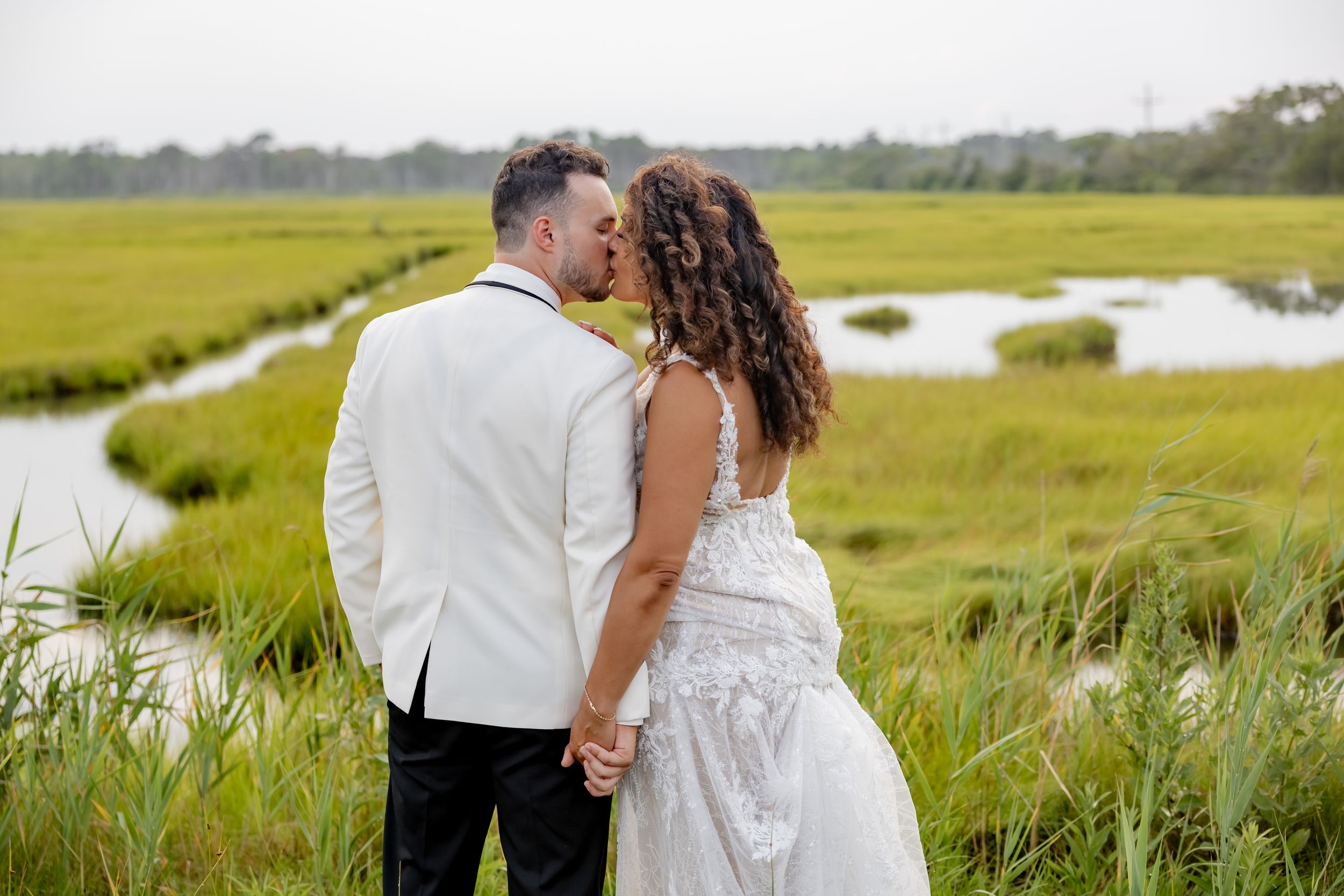 A couple shares a romantic kiss in wedding attire beside a lush green marsh waterway on their wedding day.