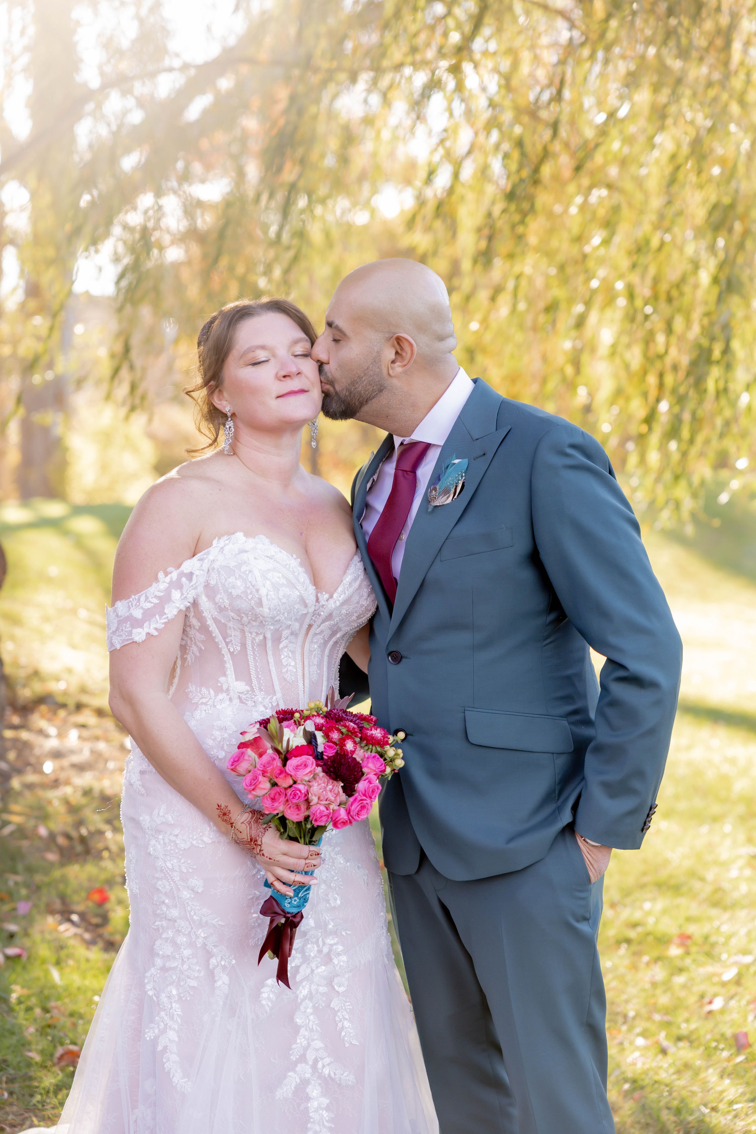 Groom in gray suit kissing bride on cheek outdoors under golden willow tree with vibrant pink bouque in Sparta, NJ.