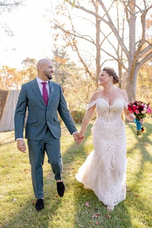 Bride and groom walking hand in hand through sunlit autumn garden, bride holding vibrant pink floral bouquet in Sparta, NJ.