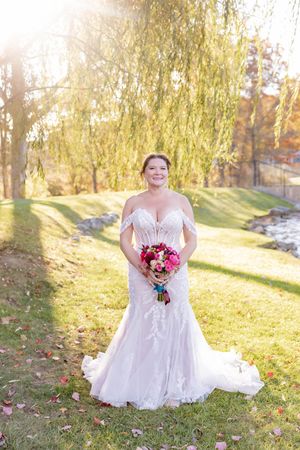 Radiant bride in off-shoulder lace gown holding vibrant pink bouquet under a golden willow tree in autumn in Sparta, NJ.