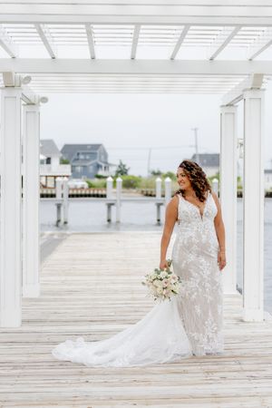 Bride in lace wedding gown holding bouquet on waterfront dock under white pergola.