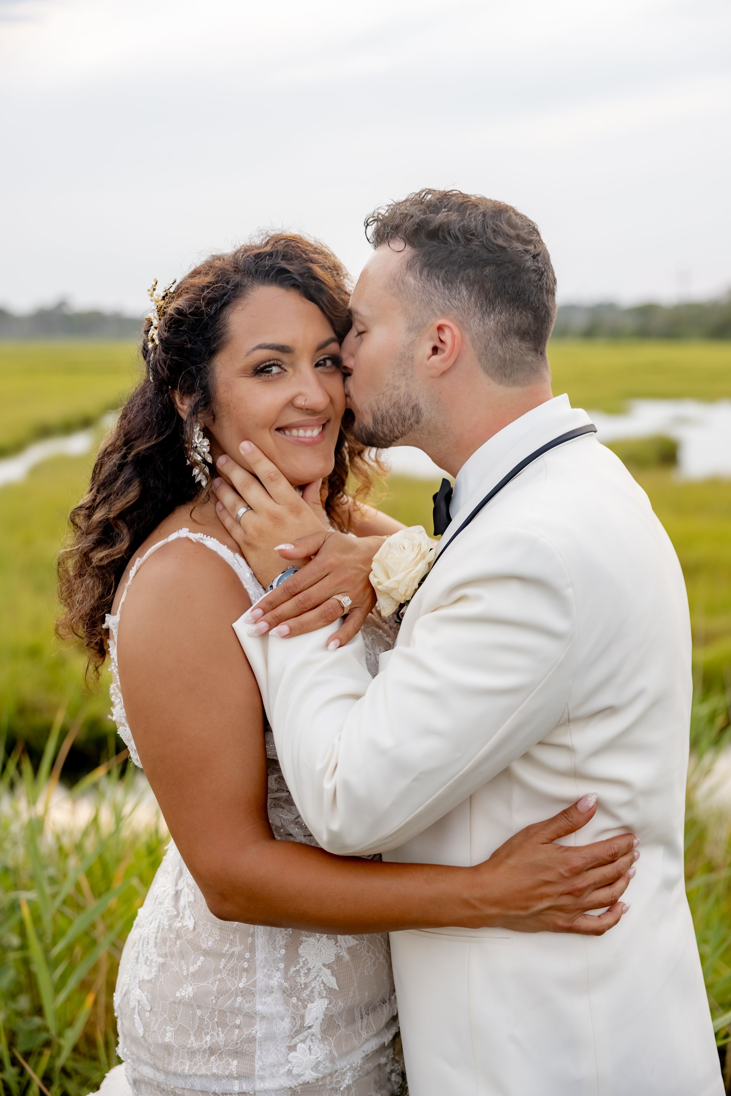 Groom in white suit kisses bride's cheek as she smiles, standing in a lush green marsh field on their wedding day.