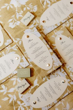 Elegant wedding stationery suite with oval menus, place cards, and gift tags arranged on floral burlap fabric.