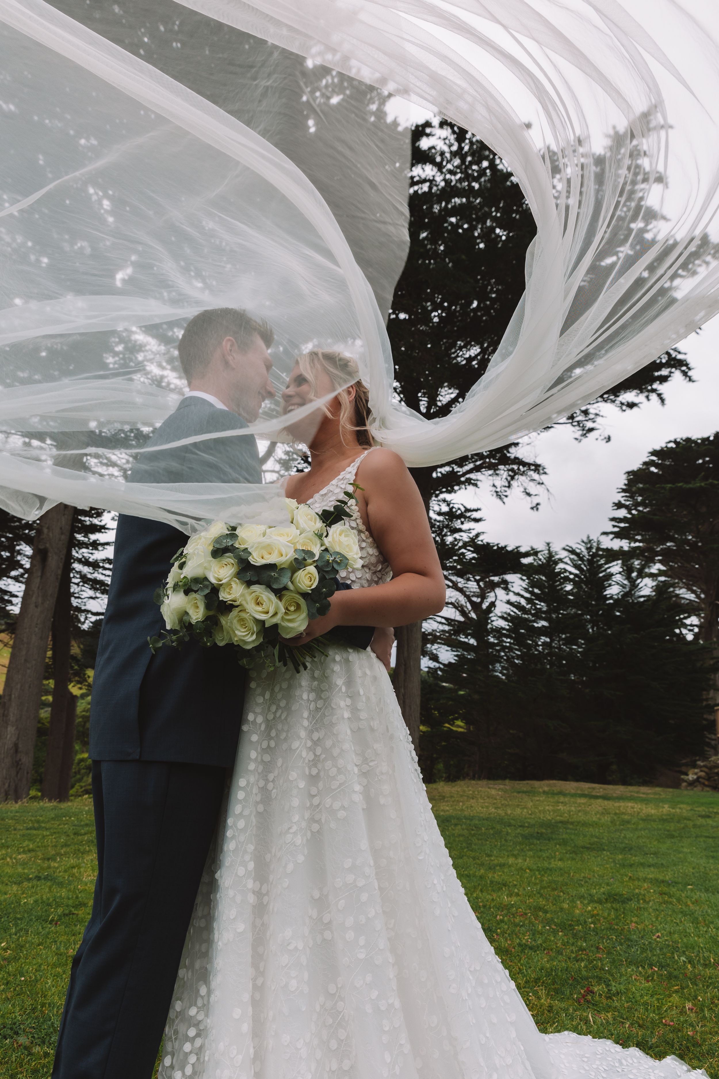 Dramatic wedding portrait with flowing veil creating artistic movement against evergreen trees.