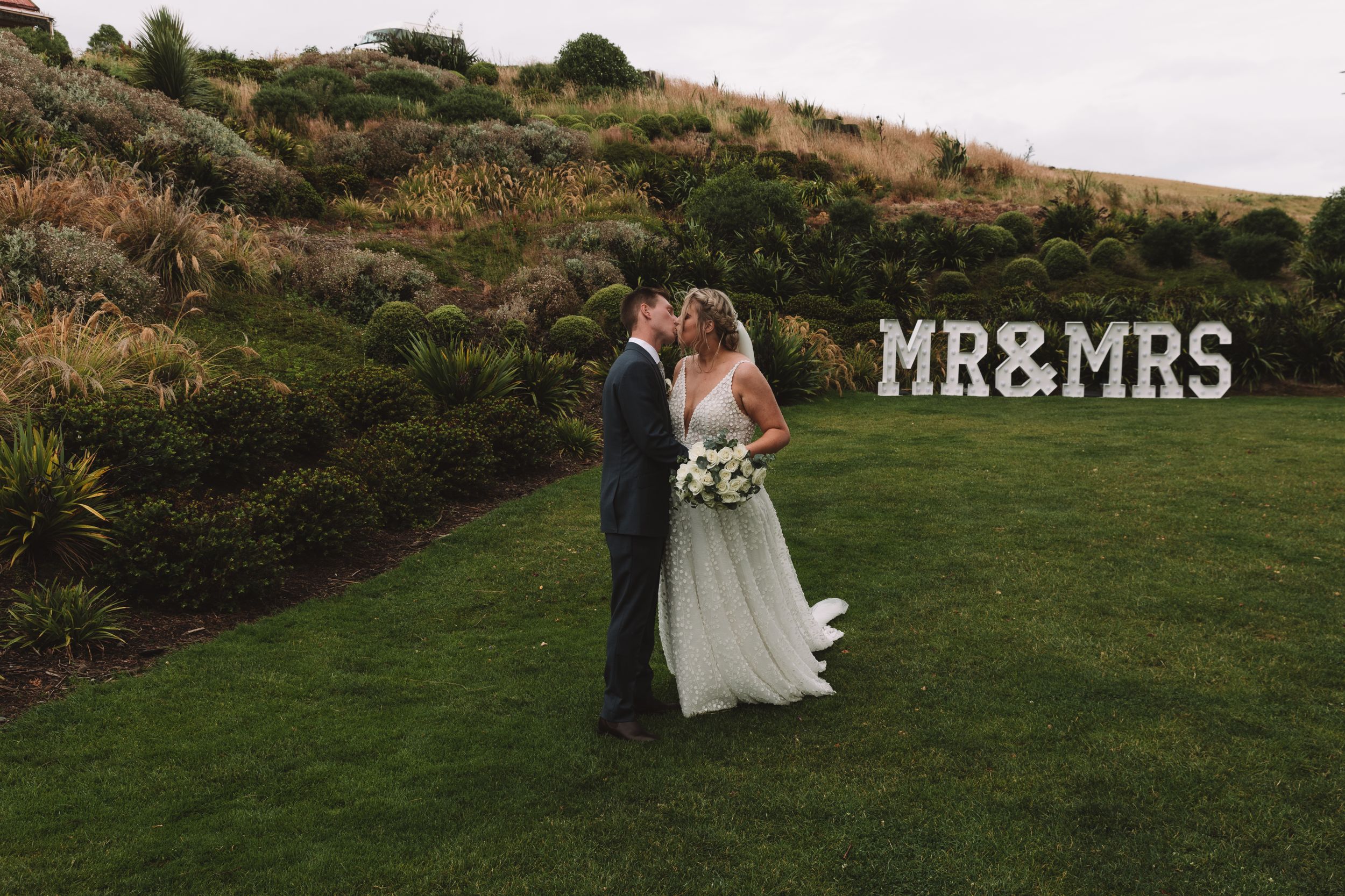 Newlyweds share an intimate moment on a hillside with MR&MRS sign in background.