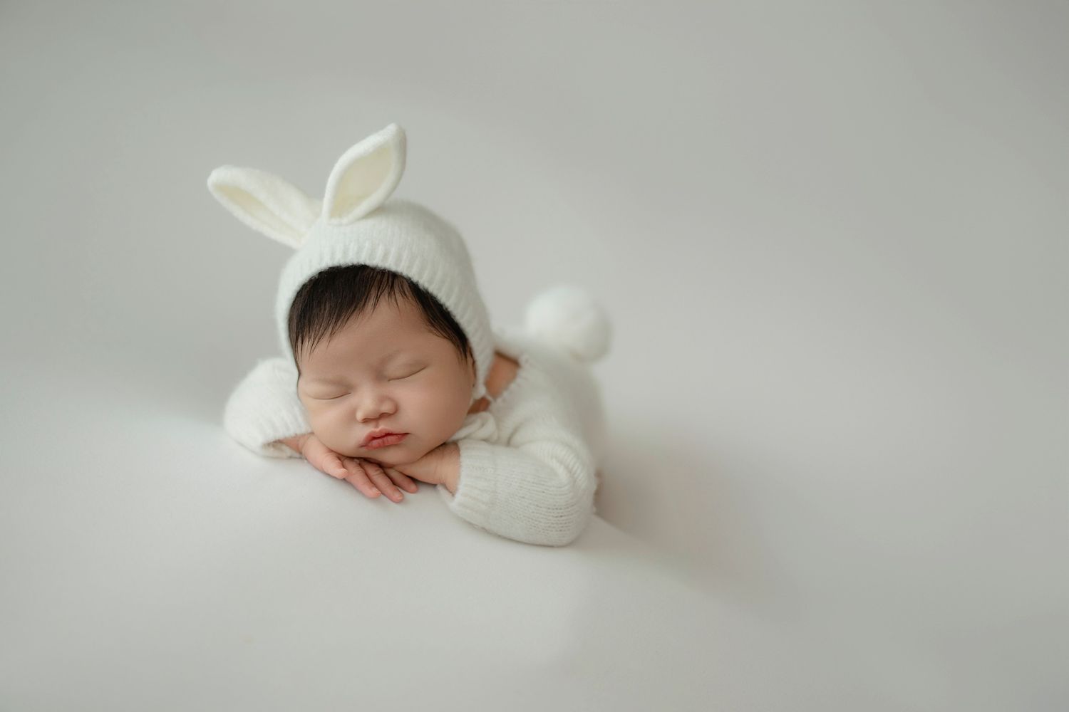 Sleeping newborn wearing white bunny-ear outfit rests peacefully on a light backdrop.