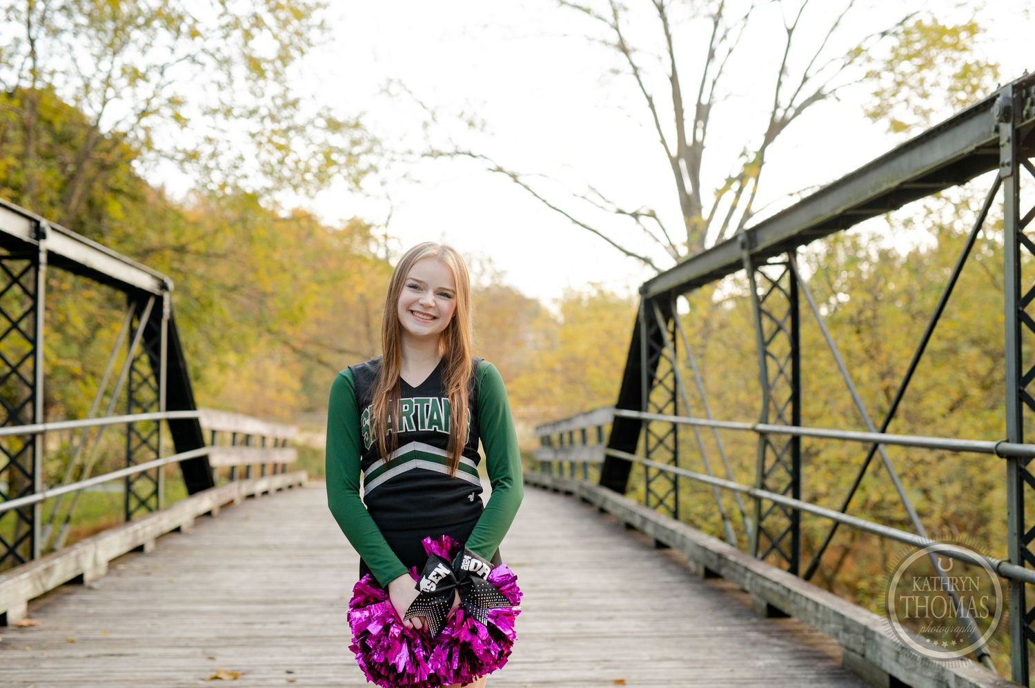 Cheerleader in green and black uniform holding pink pom poms poses on wooden bridge during autumn.