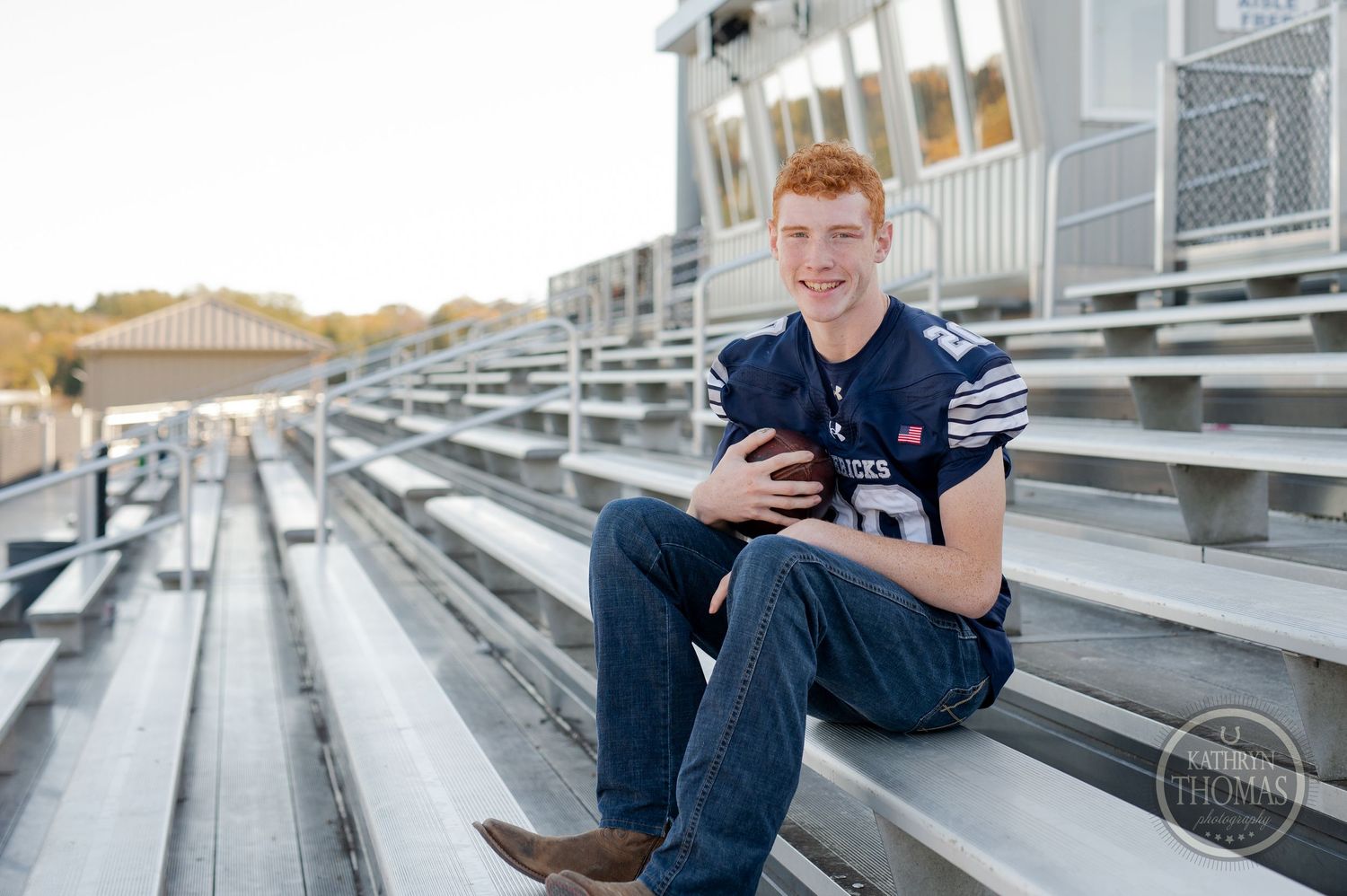 Student athlete in navy football jersey sits on stadium bleachers looking at phone.