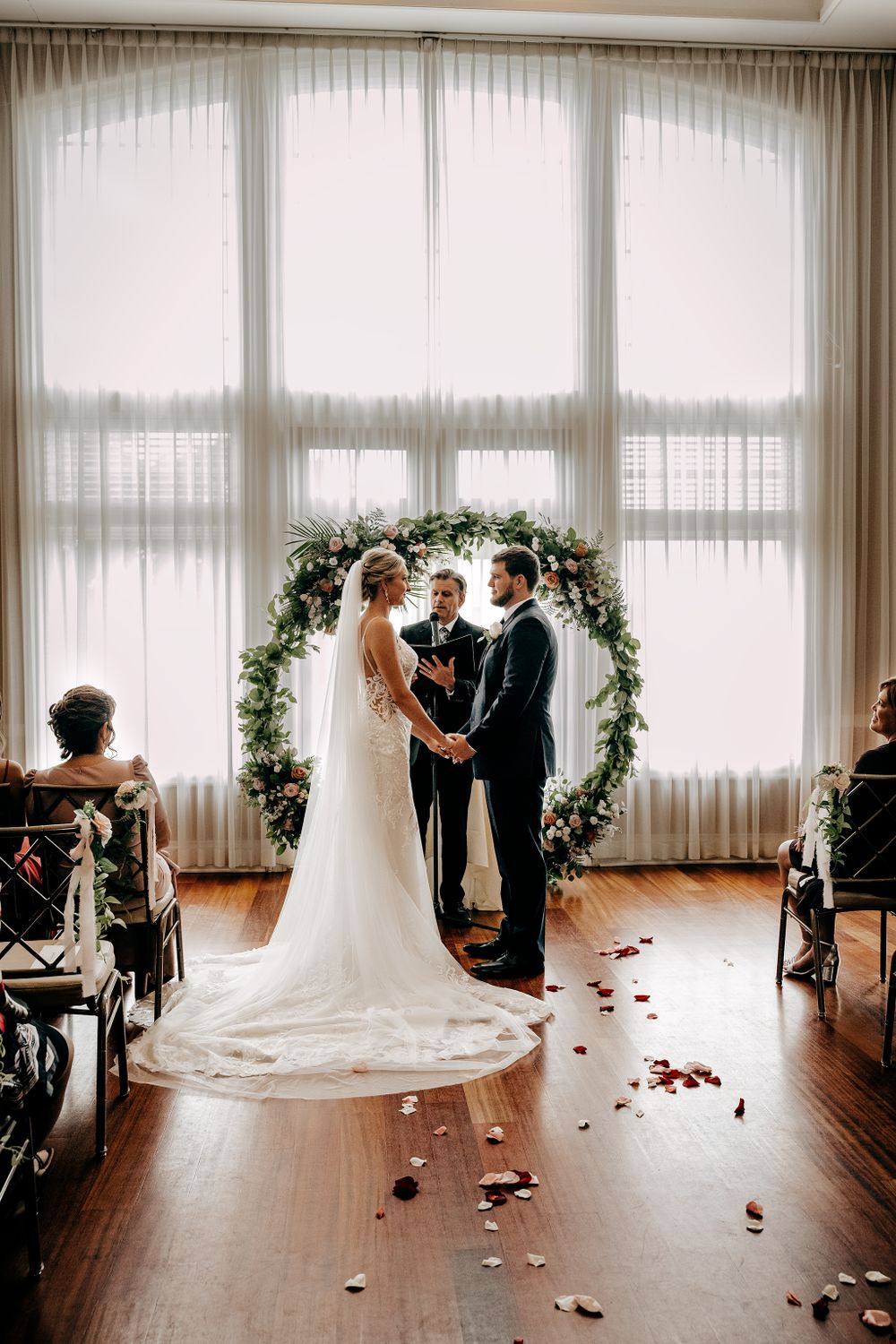 Wedding ceremony setup with floral arch against large windows and scattered rose petals.